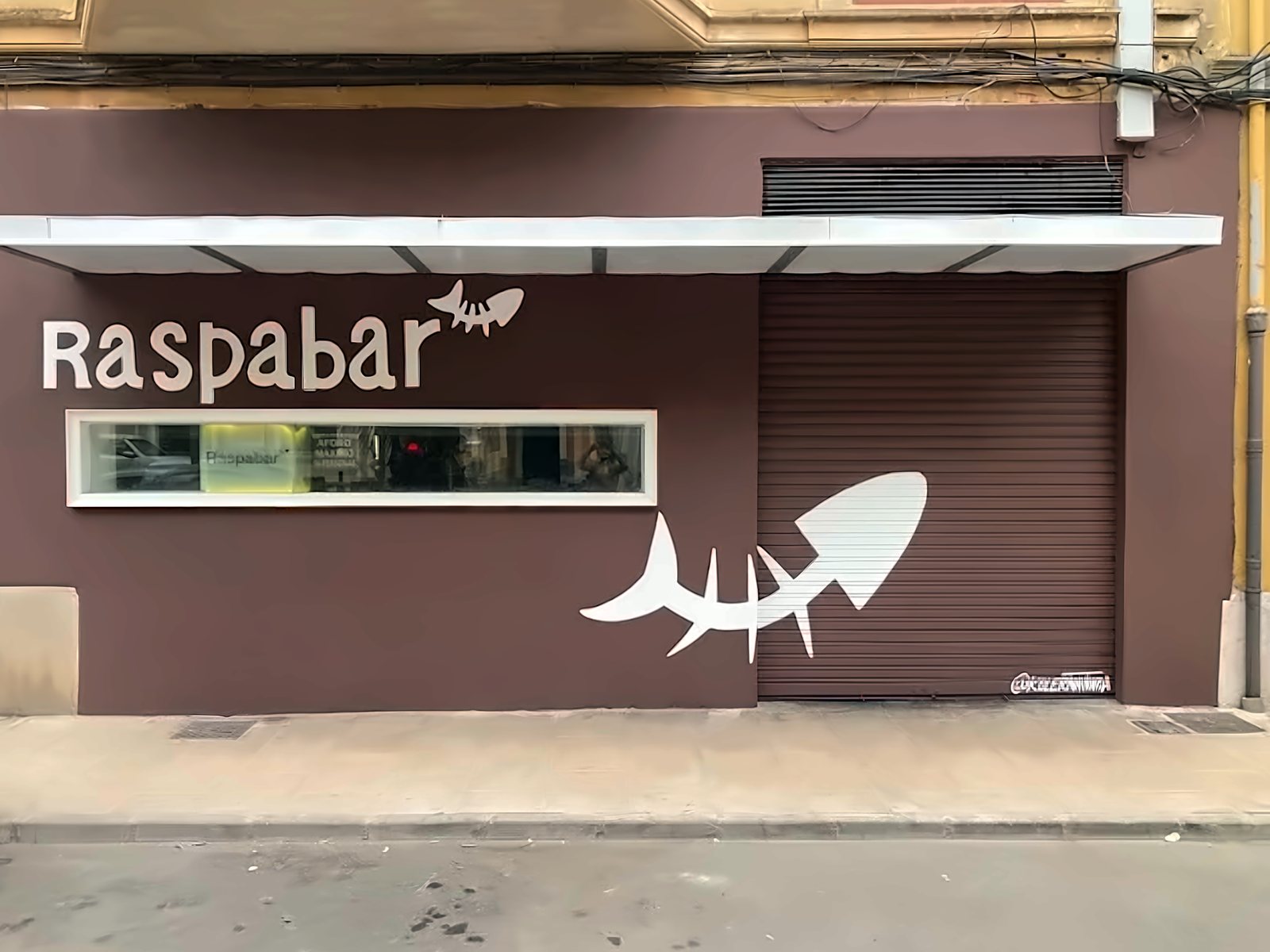 Fachada del Raspabar: pared marrón, letrero crema y la raspa de pescado pintada en el cierre